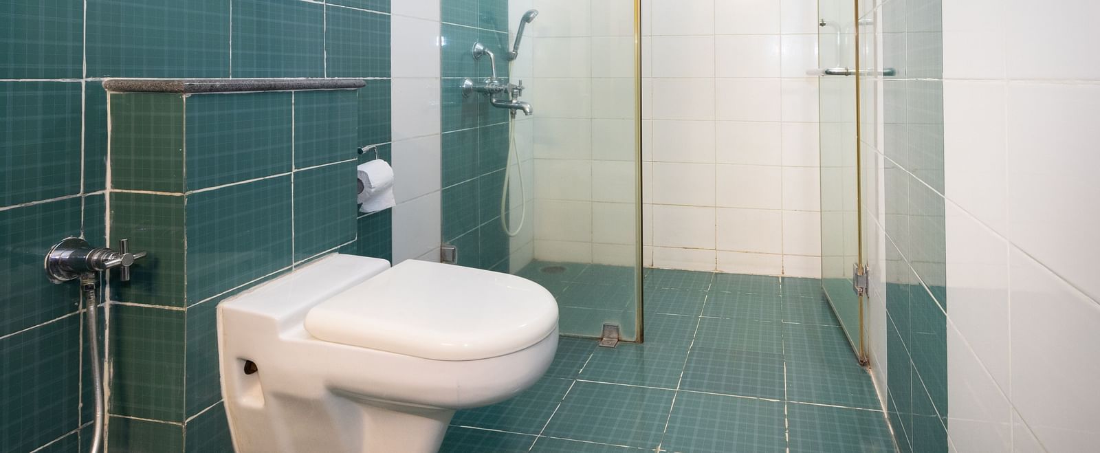 A bathroom with teal and white tiling, featuring a toilet, a shower cubicle with a glass door at Sanctum Suites Indiranagar, Bangalore.