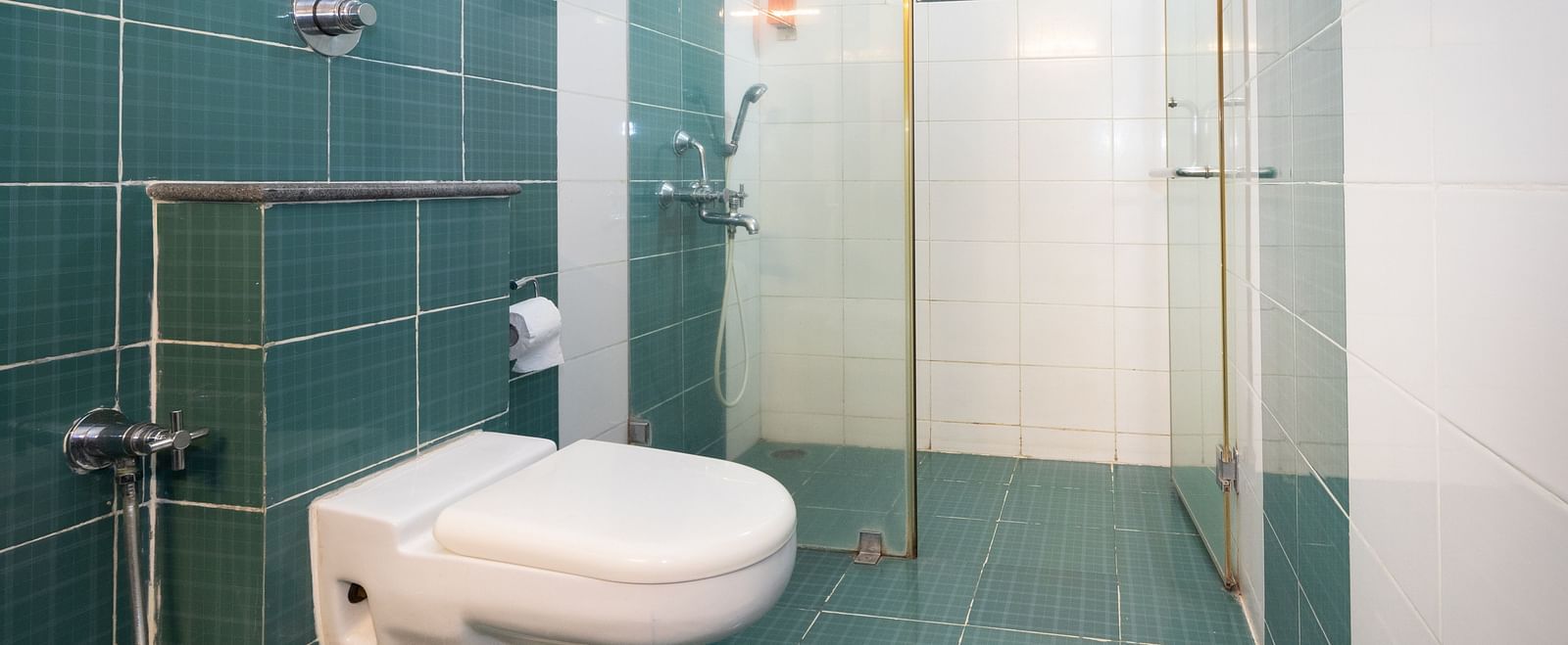 A bathroom with teal and white tiling, featuring a toilet, a shower cubicle with a glass door at Sanctum Suites Indiranagar, Bangalore.