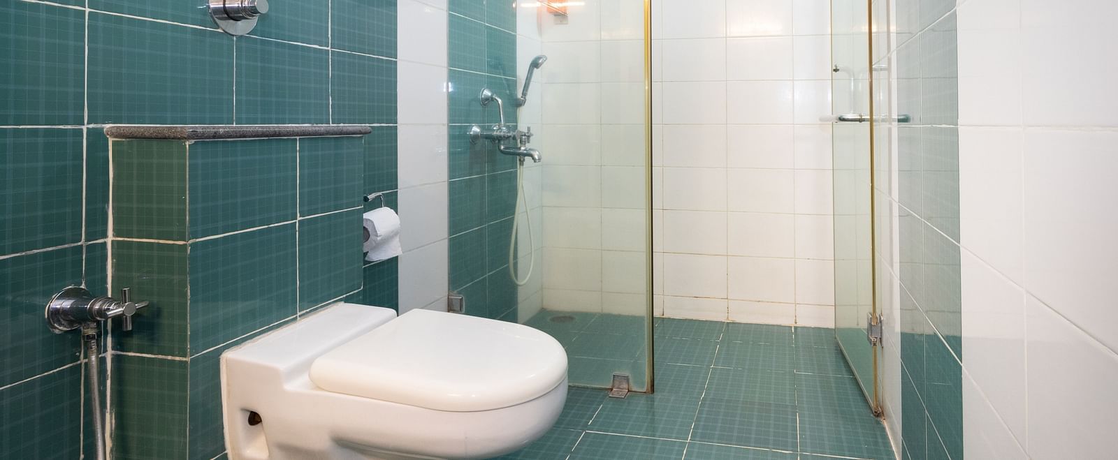 A bathroom with teal and white tiling, featuring a toilet, a shower cubicle with a glass door at Sanctum Suites Indiranagar, Bangalore.