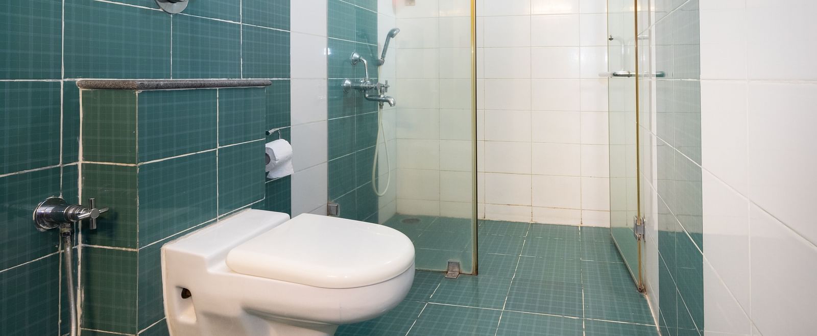 A bathroom with teal and white tiling, featuring a toilet, a shower cubicle with a glass door at Sanctum Suites Indiranagar, Bangalore.