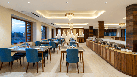 A restaurant at Tathastu Chavri Resort, Jawai featuring dining tables, chairs, a buffet counter, wooden pillars, and windows.