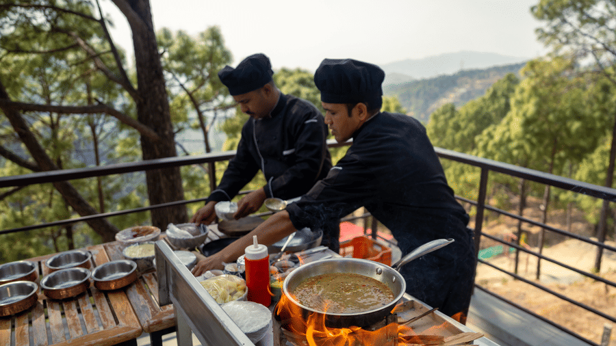 Two chefs prepare food at an outdoor cooking setup on a balcony at BluSalzz Escapade Resort, Lansdowne. Trees and hills are visible in the background.