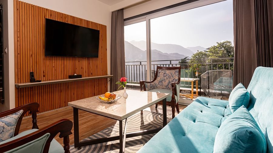 An image of a room with a flat screen TV, sofa set, centre table and an expansive glass door overlooking the mountains - The Belvedere Himalayan Retreat, McLeodganj