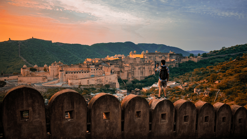A person stands on a stone battlement overlooking a vast fortress complex nestled in the hills at sunset.