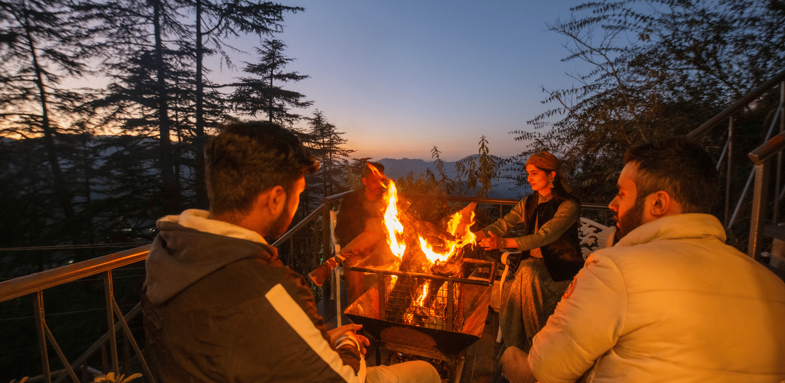 A group of guests at Perfectstayz Value Shimla (Namah Retreat) sitting around a bonfire outdoors, surrounded by pine trees and mountain views.