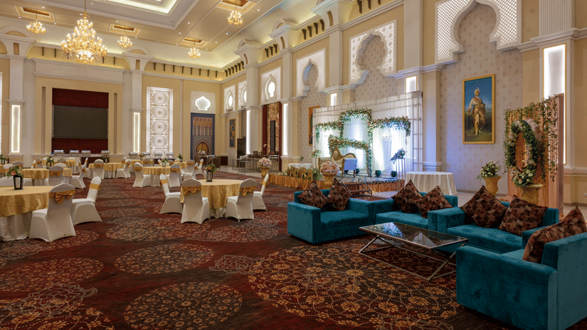 Event hall at Geeta Sarovar Portico, Panipat featuring multiple seating area is cluster setting, a well-decorated stage with flowers, and ceilings lit with lights and chandeliers hanging.