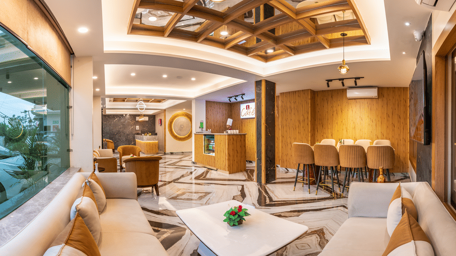 The lobby lounge at Lords Inn Katra features sofa seating, a decorative wooden ceiling panel, and large glass windows.