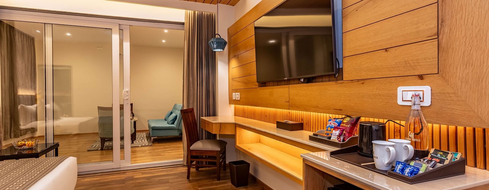An image of a Family Room with flat screen TV, modern room amenities, work desk and chair, and an expansive glass door leading to other areas of the room - The Belvedere Himalayan Retreat, McLeodganj