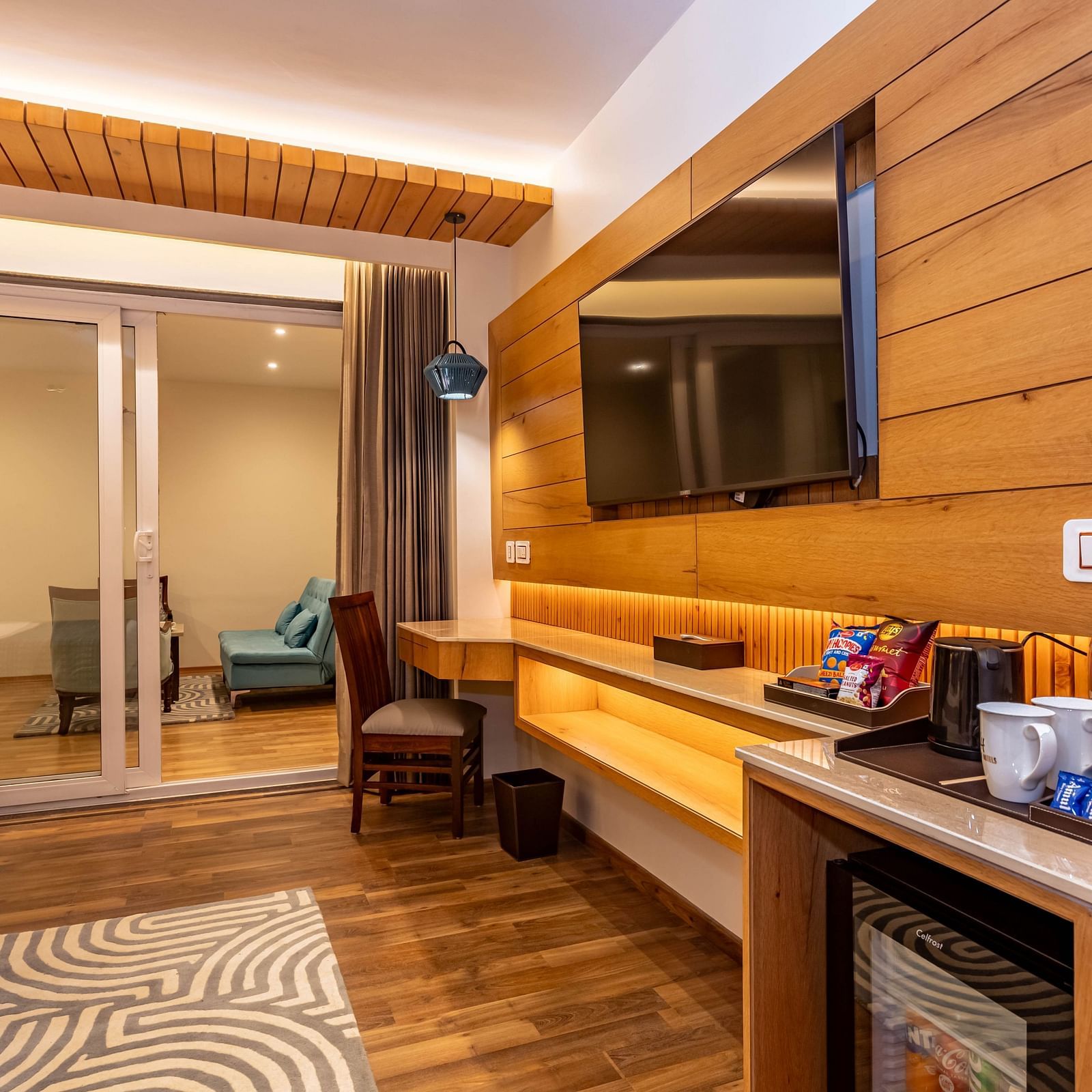 An image of a Family Room with flat screen TV, modern room amenities, work desk and chair, and an expansive glass door leading to other areas of the room - The Belvedere Himalayan Retreat, McLeodganj