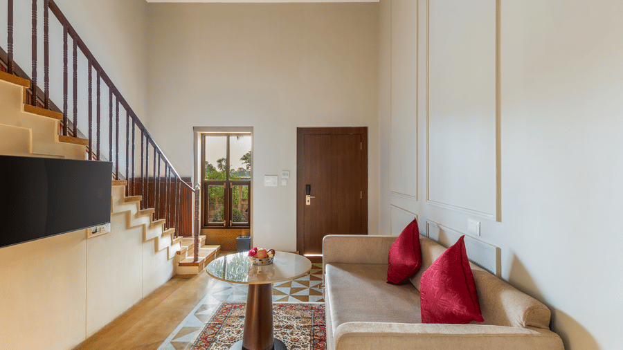 Living area in a suite at EsthereaRaj Leela, Ranakpur, with a sofa, red cushions, a coffee table, and wooden stairs leading to an upper level.