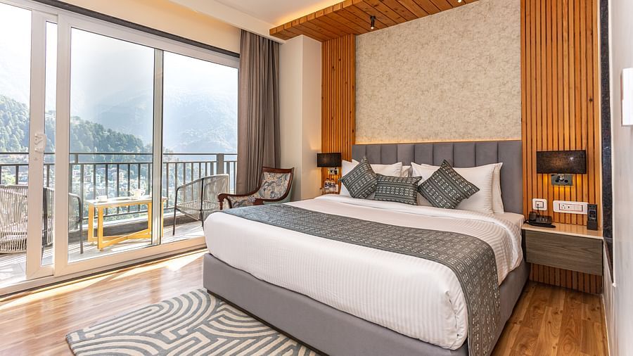 An image of a Suite with a bed, bed side table, lamp shades, and an expansive glass door leading to the balcony - The Belvedere Himalayan Retreat, McLeodganj