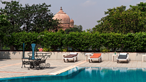 A bright blue swimming pool is shown with sun loungers and a large, domed monument visible in the background - Clarks Avadh, Lucknow