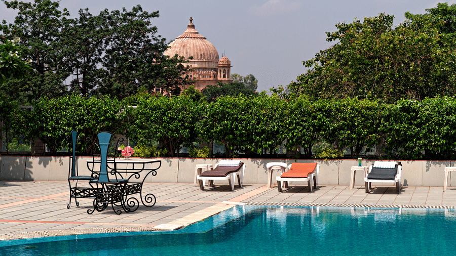A bright blue swimming pool is shown with sun loungers and a large, domed monument visible in the background - Clarks Avadh, Lucknow