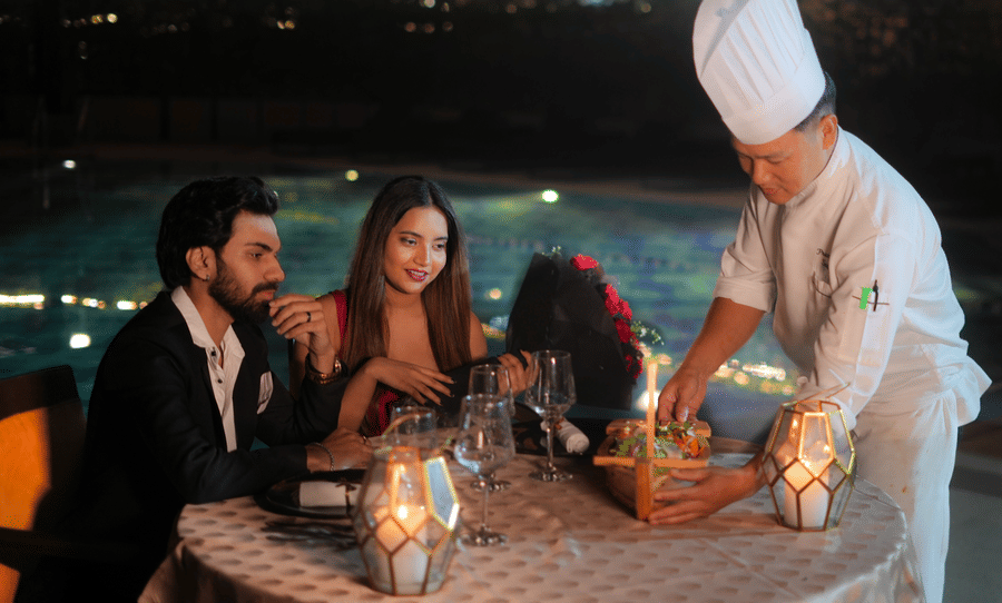 A chef serving dishes to a man and a woman seated at a table beside a swimming pool at The Suryaa, New Delhi.