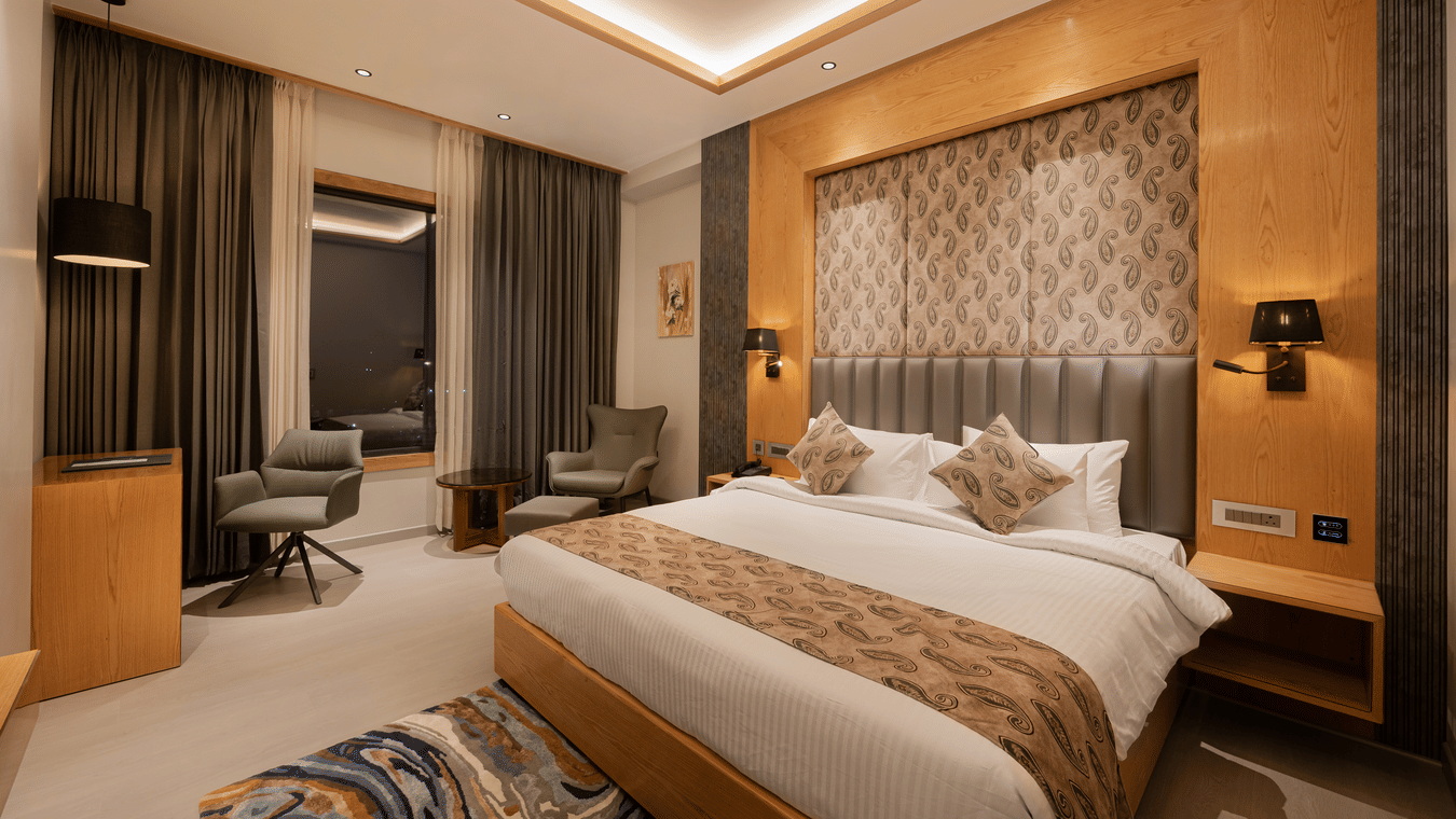 A bright guest room with a king-sized bed, decorative headboard, and warm wooden wall panelling at Ananta Express, Katra.