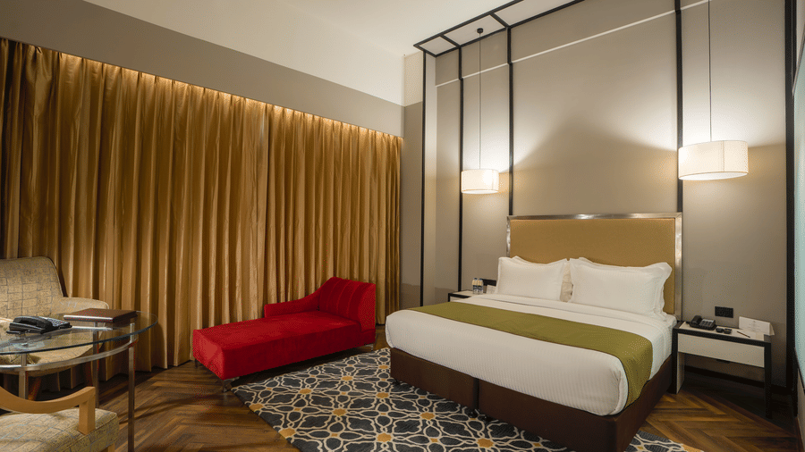 Guest room at Polo Hotel, Agartala with a king bed, gold curtains, a red chaise lounge, pendant lights and a patterned rug on a herringbone wood floor.
