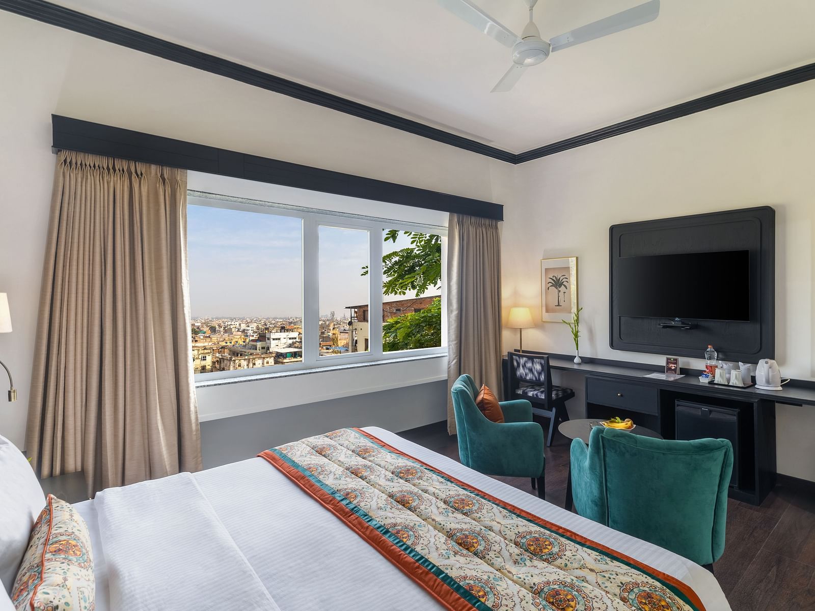 a spacious room featuring a king size bed, two green armchairs, a Television cabinet with a table lamp and a window overlooking the city - Hotel Rajdarshan, Udaipur.