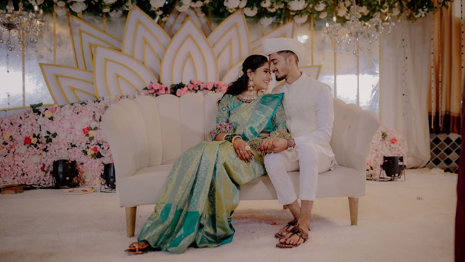 Bride in saree and groom in  sherwani sit on a love seat before a backdrop of lotus petals and flowers at their wedding reception - The Grand Legacy Resort and Spa