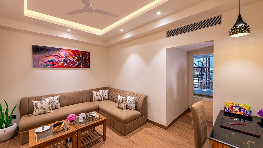 Living area with a sectional sofa, a coffee table, and a view into an adjoining space. Features a distinctive ceiling design, a ceiling fan, and a colourful abstract painting on the wall - Lords Eco Inn Dehradun.