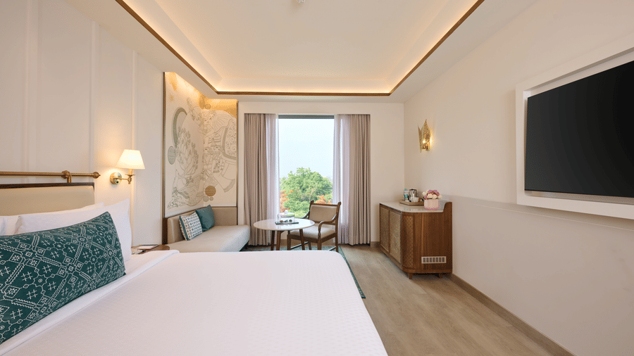 Standard Room view at Prachi Sarovar Premiere, Bhubaneswar, featuring a bed, a wall television, a cabinet, a small sofa, and a window.