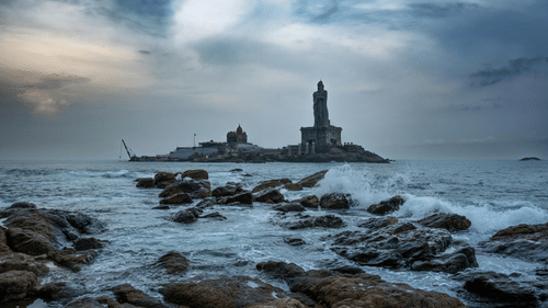 A rocky shoreline in the foreground leads to a distant island with a building and a statue of Thiruvalluvar silhouetted against a cloudy sky.