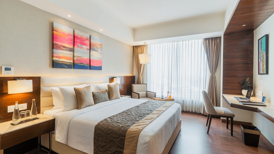 A modern bedroom at Nirvana Sarovar Portico, Gorakhpur featuring a king-size bed, abstract wall art, study desk, and large window.