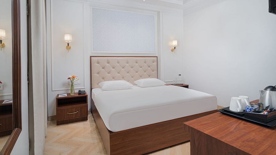 A spacious bed featuring a cushioned headboard, with bedside tables on either side, and a wooden table holding a tray with a kettle and cups | Hotel Ganga Maiya
