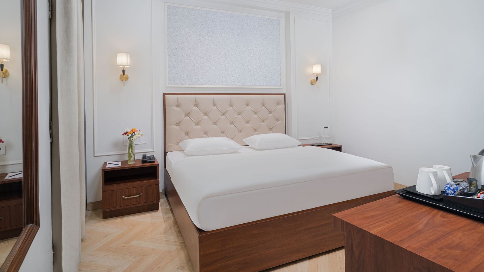 A spacious bed featuring a cushioned headboard, with bedside tables on either side, and a wooden table holding a tray with a kettle and cups | Hotel Ganga Maiya