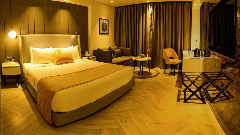 A spacious hotel room with a king-sized bed, a seating area, a work desk, and warm golden lighting creating an inviting ambiance. - Hotel Kanha Shyam, Prayagraj