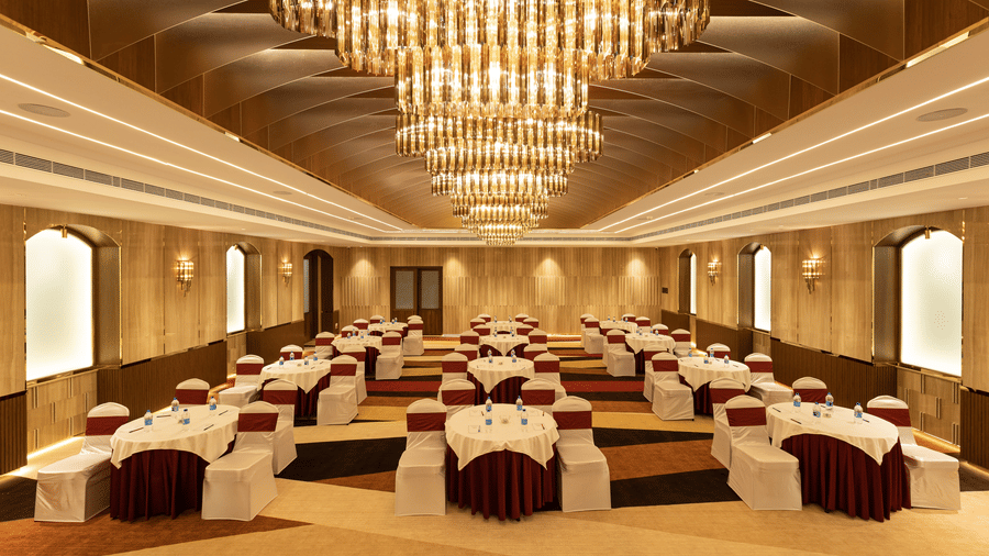 A luxury banquet hall arranged with round dining tables and elegant maroon accents under large crystal chandeliers - Jenneys Residency, Coimbatore