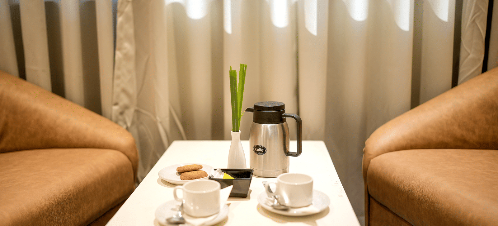 A close-up of a small table set for tea or coffee between two tan armchairs by a window at Pravasa Hotel, Kolhapur.