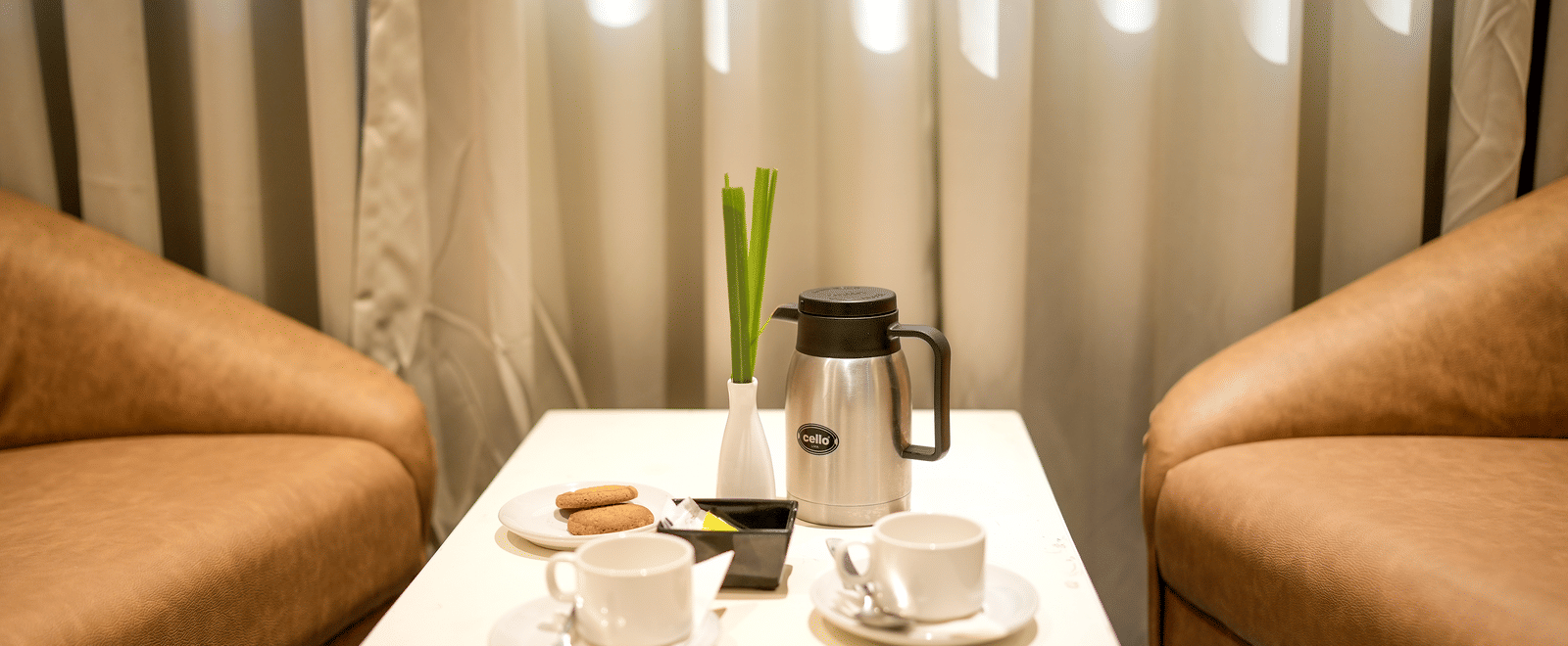 A close-up of a small table set for tea or coffee between two tan armchairs by a window at Pravasa Hotel, Kolhapur.