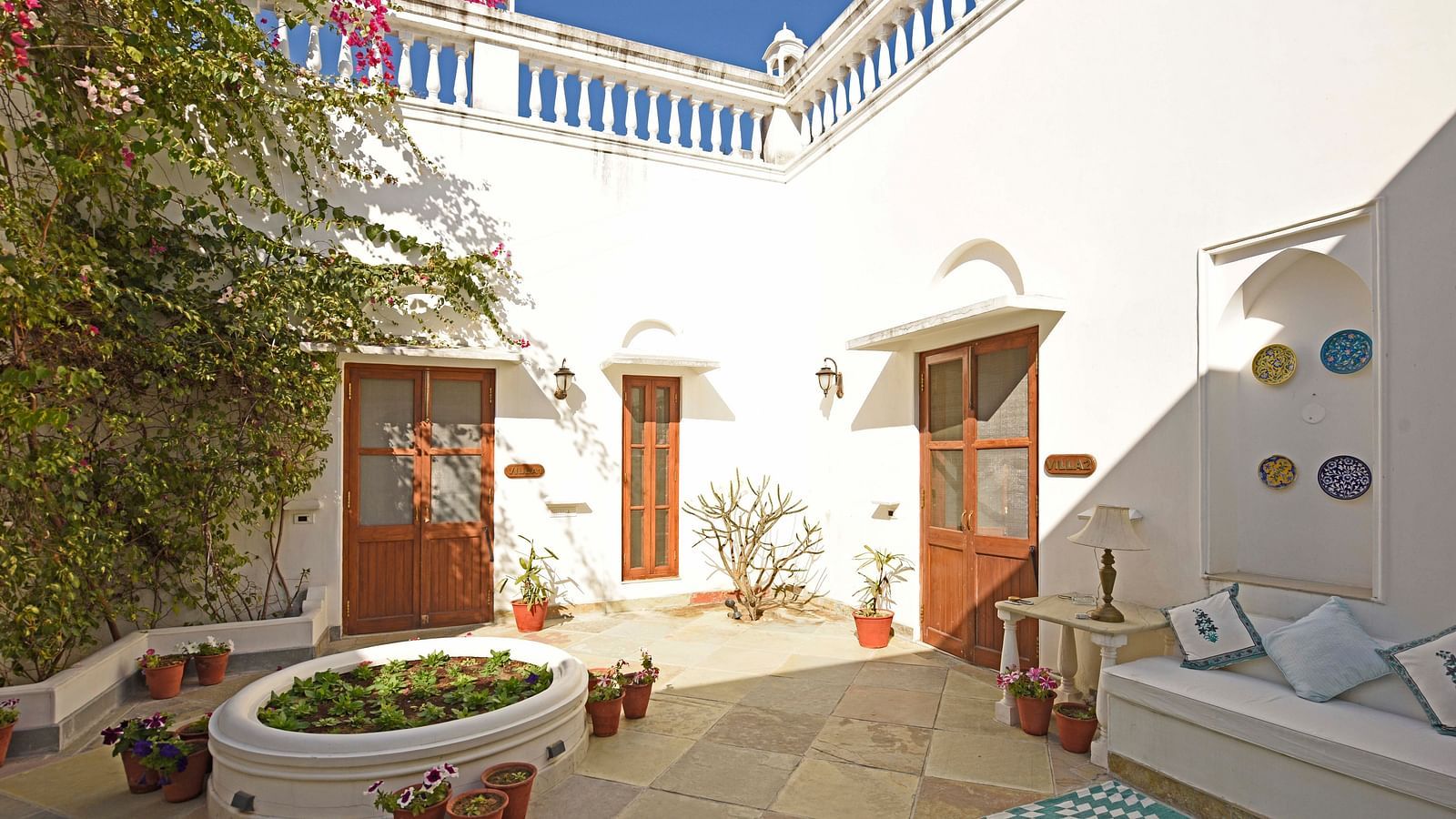 A bright courtyard with white walls, featuring wooden doors, potted plants, a built-in planter, and a seating area with cushions- Shahpura Bagh.