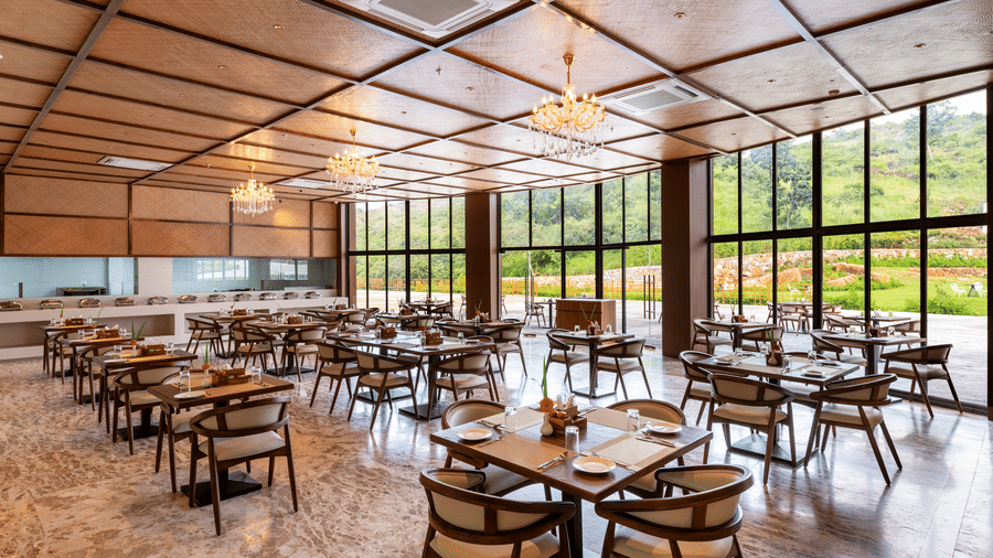 A side angle view of Lepra at Osian Valley Resort & Spa featuring a grid of wooden tables and chairs with white cushions. 