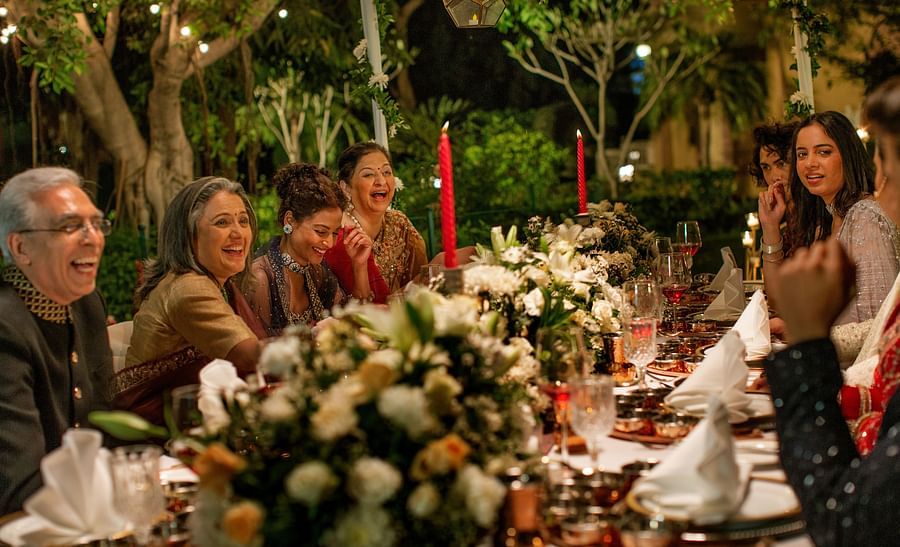 An outdoor evening dinner party with people laughing around a table decorated with candles and floral arrangements, strung lights above adding a warm glow.
