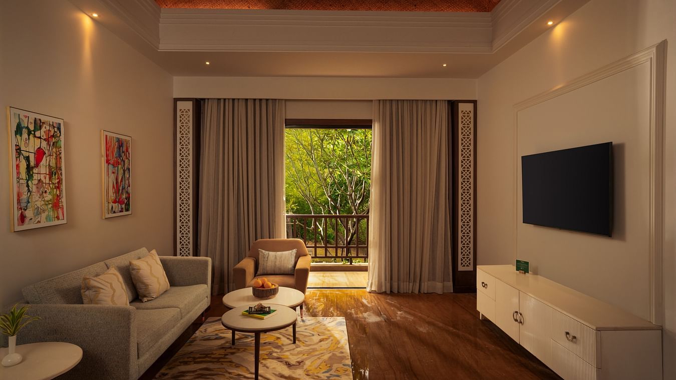A side angle of spacious Grand Suite Living Room at The Ananta Udaipur with comfortable seating, a TV, and large balcony with a jungle view overlooking the outdoors, a flat screen TV,, sofa dn a tea table .