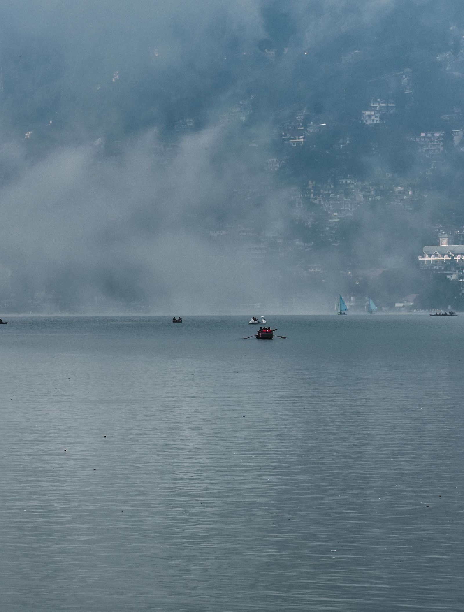 A view of a large, misty lake with several small boats on the water, and hills with buildings in the background.