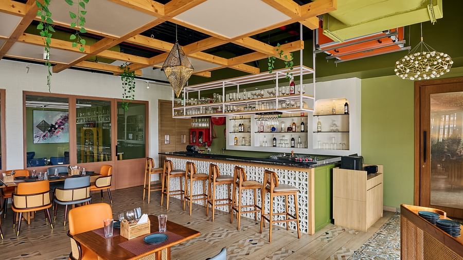 A brightly lit, modern restaurant interior featuring a bar area with wooden stools, several small dining tables with colourful chairs, and a unique ceiling design with wooden beams and hanging plants - Voyage The Solitaire Bliss.