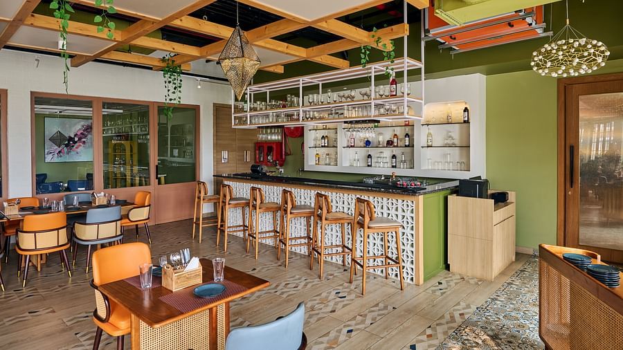 A brightly lit, modern restaurant interior featuring a bar area with wooden stools, several small dining tables with colourful chairs, and a unique ceiling design with wooden beams and hanging plants - Voyage The Solitaire Bliss.