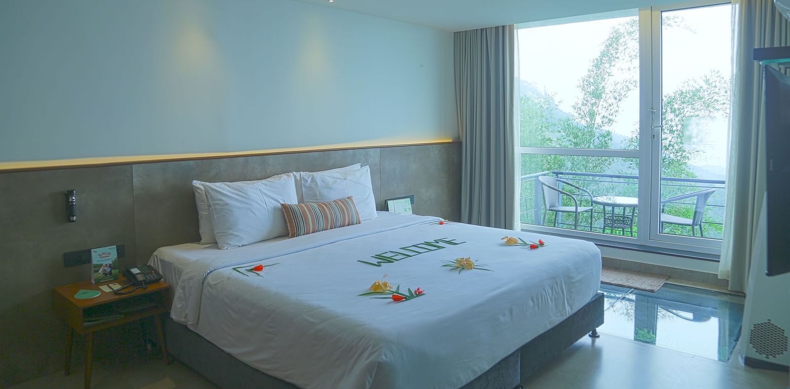 An image of the panorama view room with a king size bed, an expansive glass door leading to the outdoor space with chair and table - Great Trails Yercaud by GRT Hotels