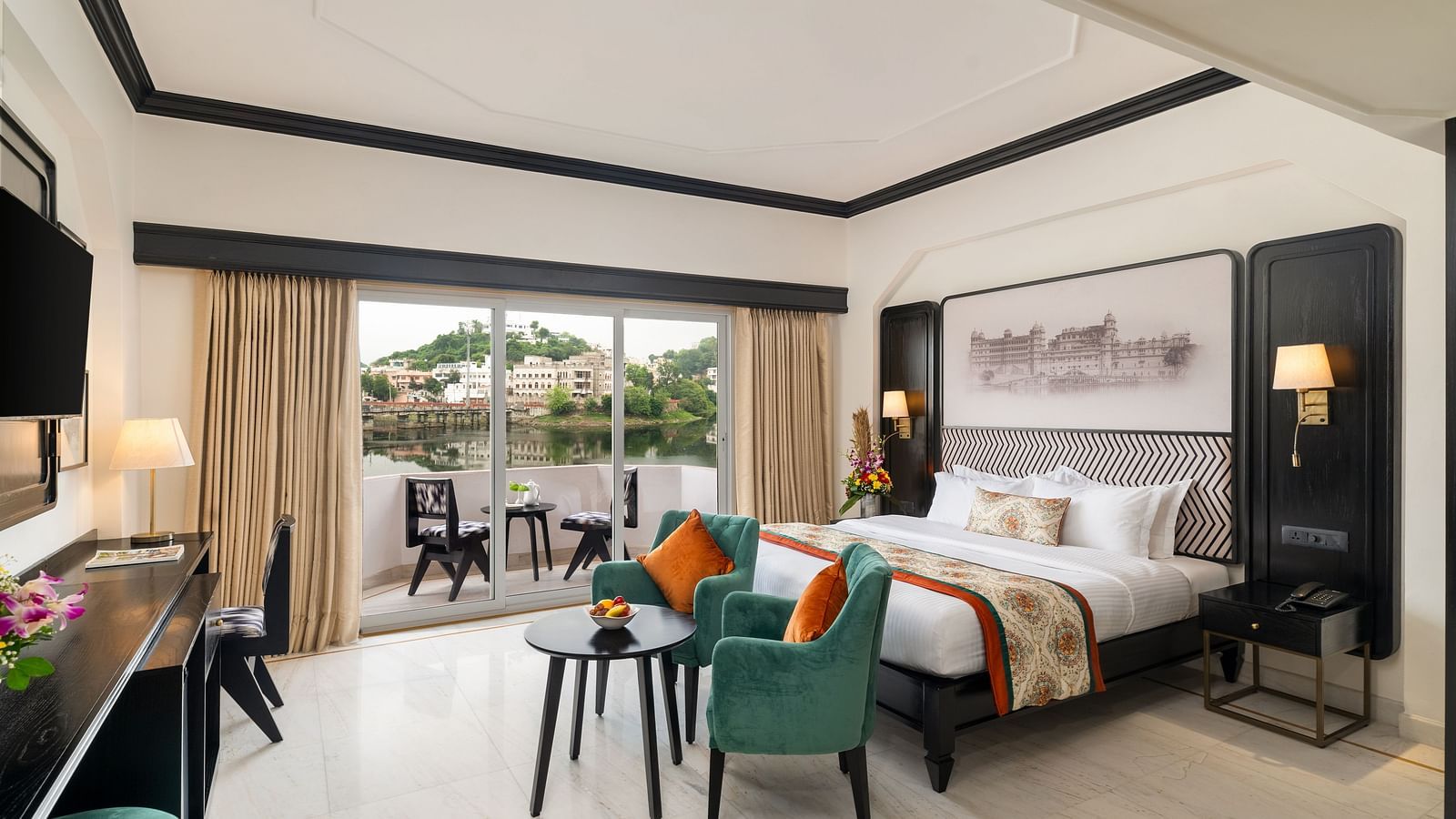 A spacious room with balcony featuring a king size bed, two  comfortable green chairs, a cabinet with a table lamps - Hotel Rajdarshan, Udaipur.