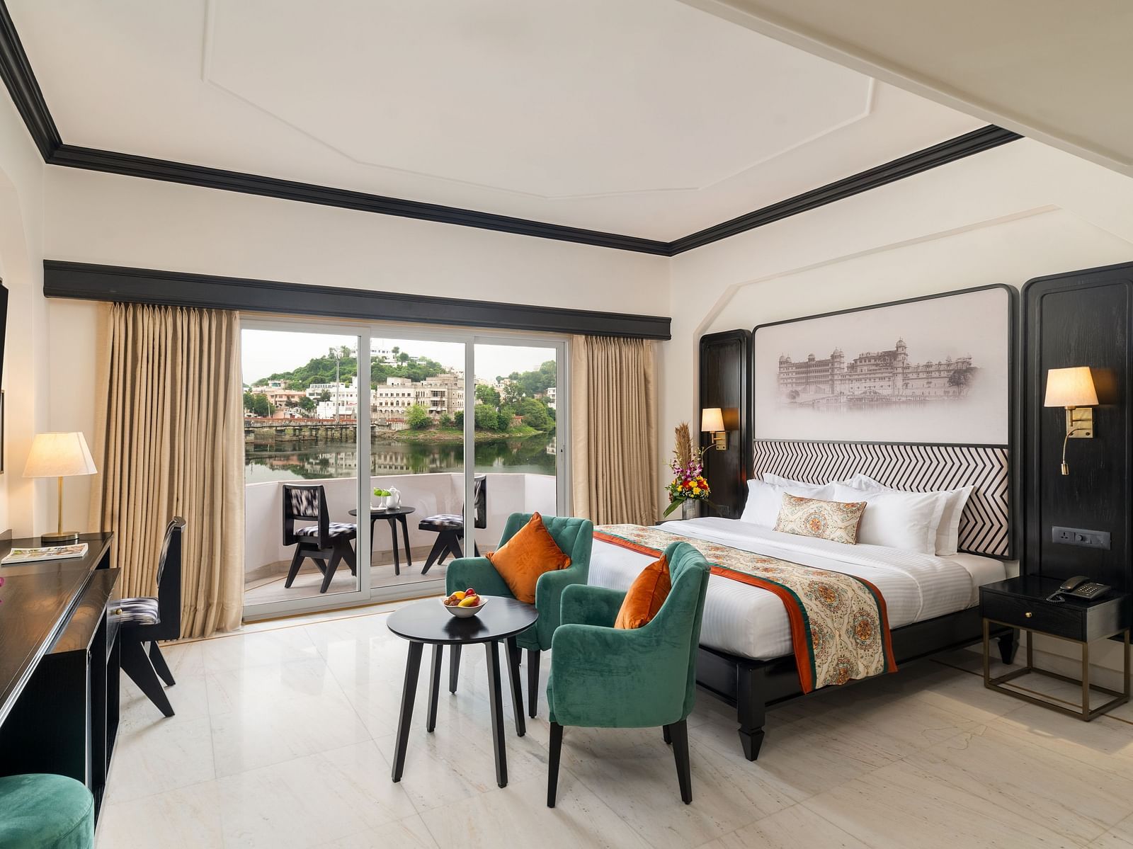 A spacious room with balcony featuring a king size bed, two  comfortable green chairs, a cabinet with a table lamps - Hotel Rajdarshan, Udaipur.