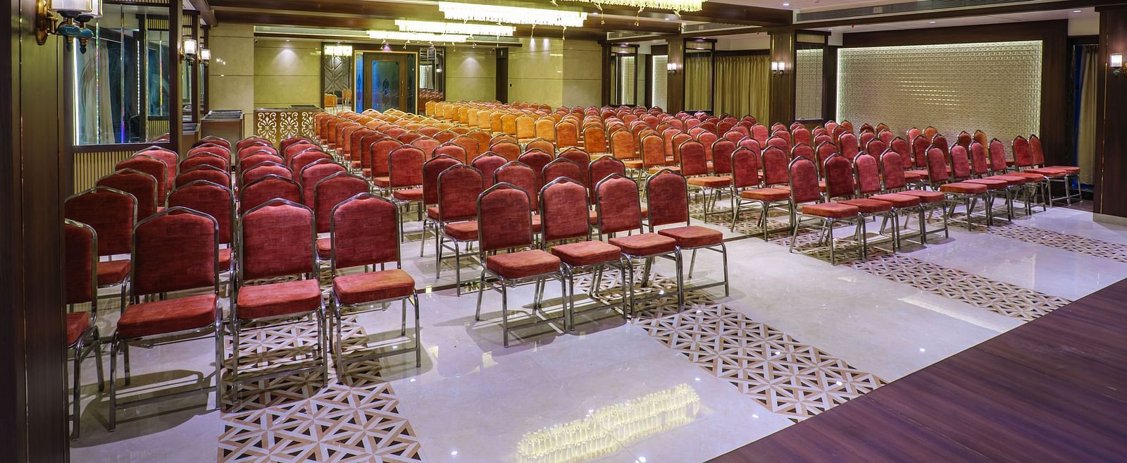 A large room is set up with rows of red chairs facing a stage area with warm lighting - Melior Inn