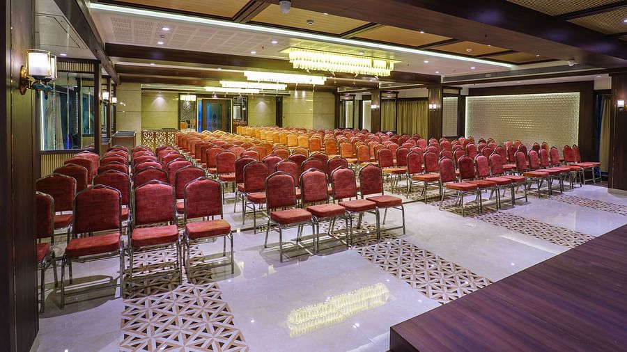 A large room is set up with rows of red chairs facing a stage area with warm lighting - Melior Inn