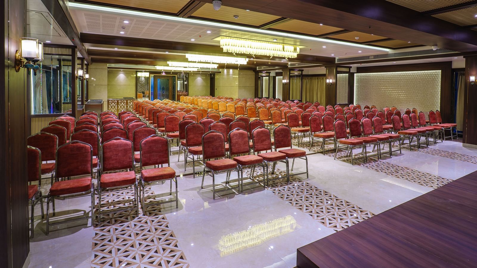A large room is set up with rows of red chairs facing a stage area with warm lighting - Melior Inn