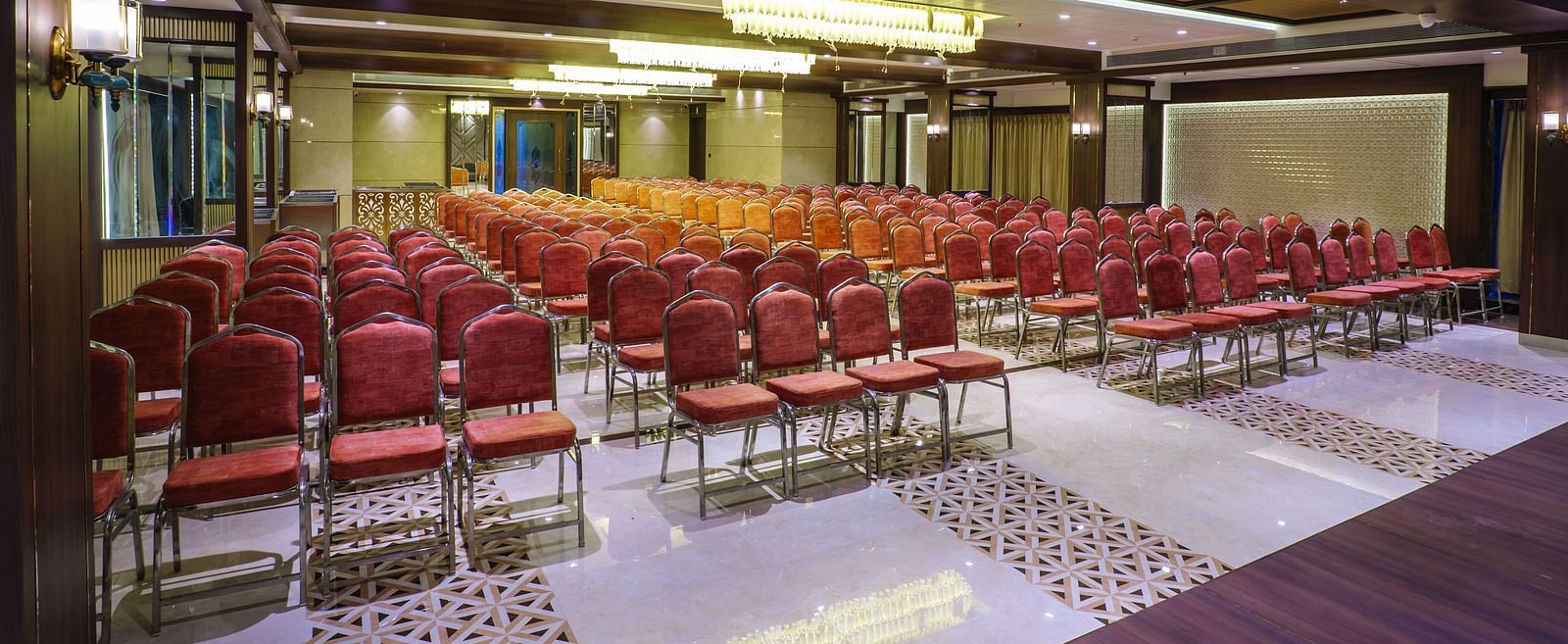 A large room is set up with rows of red chairs facing a stage area with warm lighting - Melior Inn