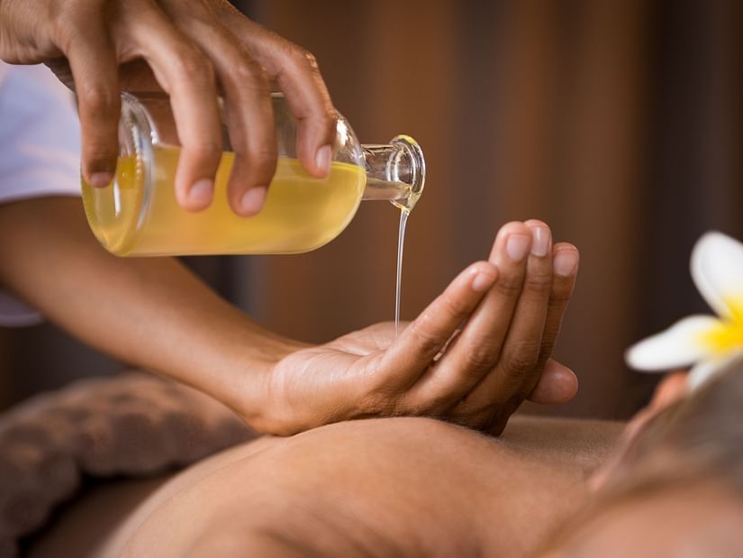 A person pours a liquid from a bottle onto their hand in a spa setting, with a flower nearby