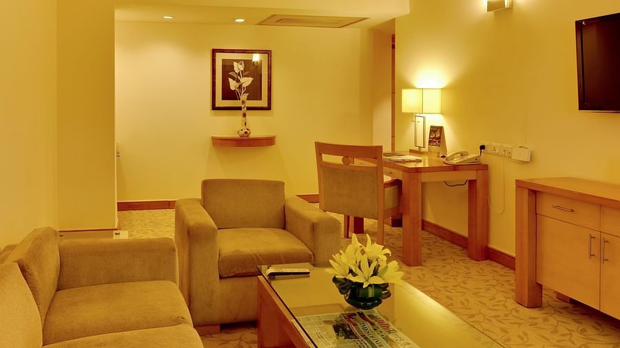 An image of a seating area with sofa sets, centre table, flat TV screen, a cabinet, and a work desk with chair in a Deluxe Suite - The Suryaa New Delhi