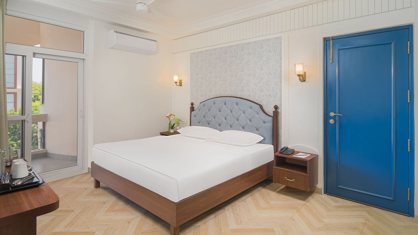 Side view of a spacious room with a large bed featuring a cushioned headboard, warm and inviting lighting, and bedside tables on either side | Hotel Ganga Maiya
