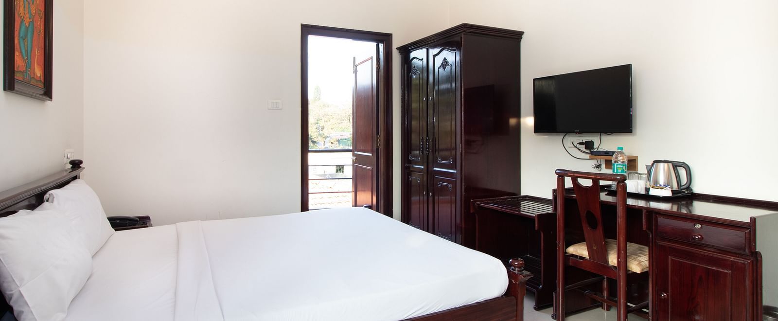 A bright room with a double bed with white linens, complemented by dark wooden furniture and a doorway leading to the balcony at Sanctum Suites Indiranagar, Bangalore.